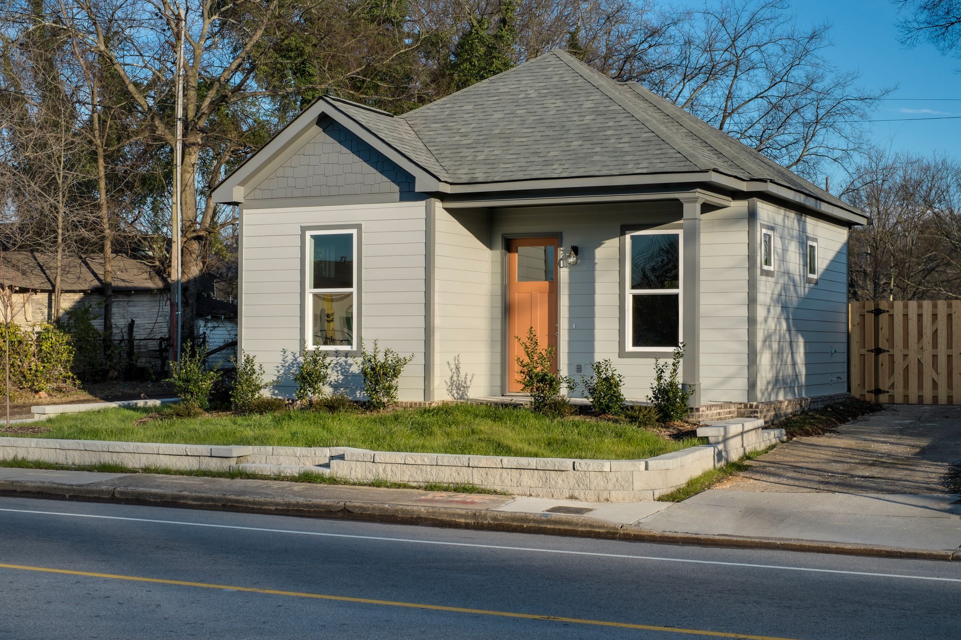 Small gray rental home with a light brown door and two front windows. There are small shrubs in front and a low stone wall.