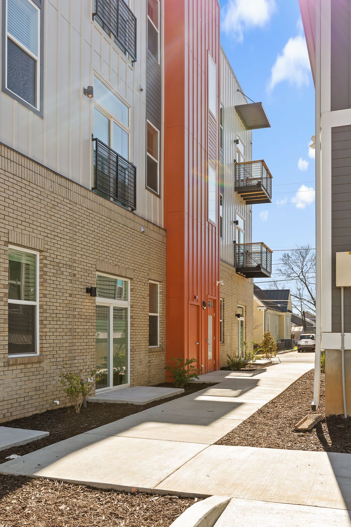 Sidewalk between multi-story buildings, brick, orange panels, balconies, and blue sky.
