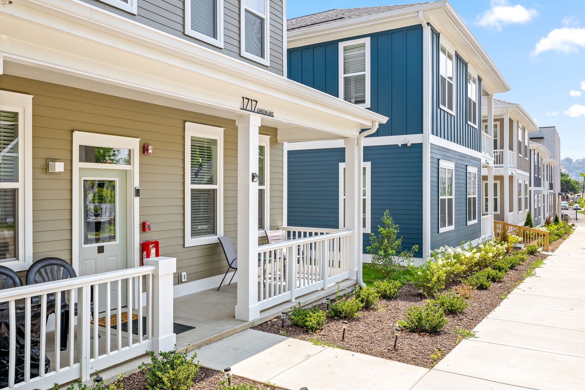 Multi-story quadplexes with front porches and landscaping, featuring tan, gray, and blue siding, with a sidewalk running along the front.