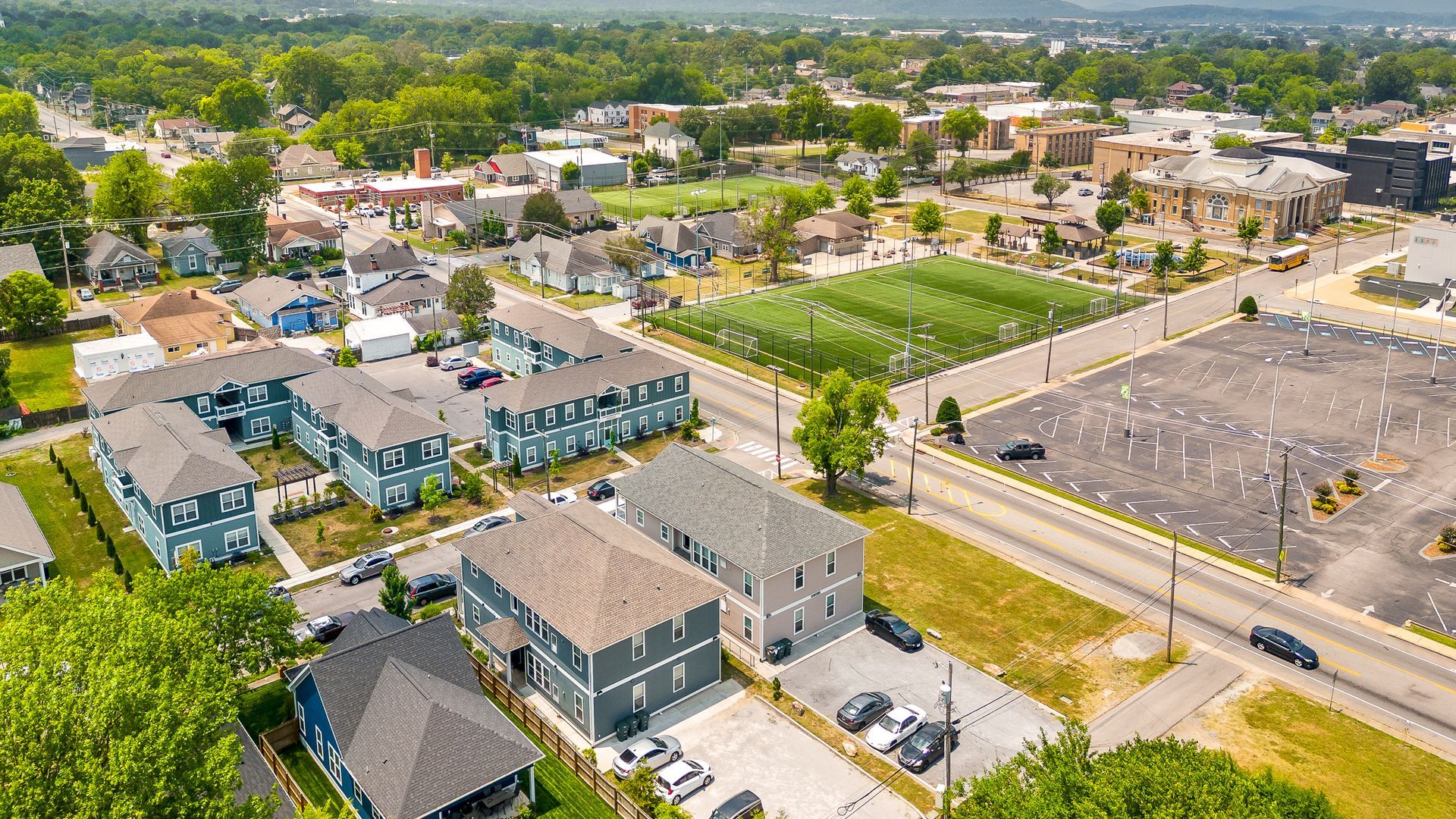 Aerial view of a town with houses, a field, and a parking lot. Buildings are blue and gray.