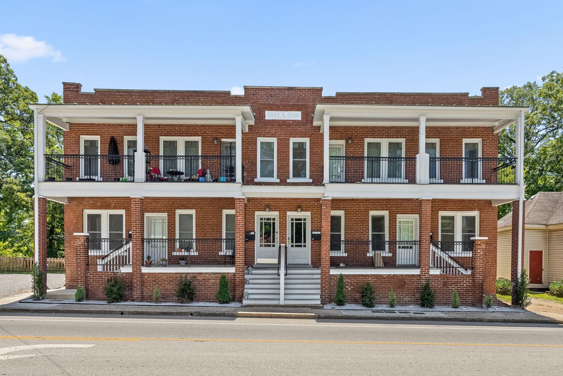Two-story brick apartment building with balconies, fronted by a street and trees.