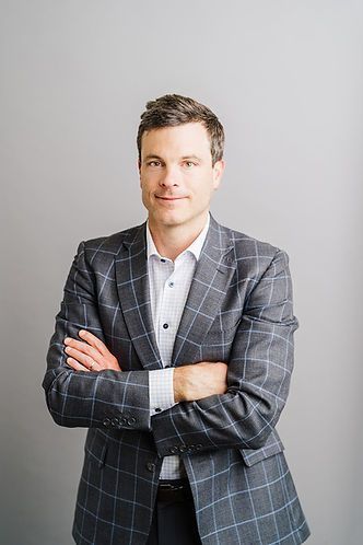 Man in a checked blazer with arms crossed, smiling, against a gray backdrop.