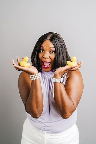 Woman with dark hair and a surprised expression, holding two yellow rubber ducks in front of a gray background.