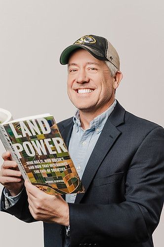 A smiling man wearing a baseball cap and suit jacket holds a book titled 