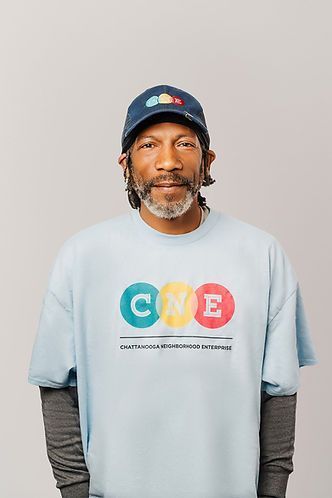 Man with graying hair and beard wearing a blue hat and light blue t-shirt with a colorful logo; standing against a light gray background.