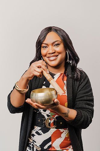 Woman with a beaming smile holding a singing bowl and striker. She is wearing a colorful dress and dark cardigan against a white background.