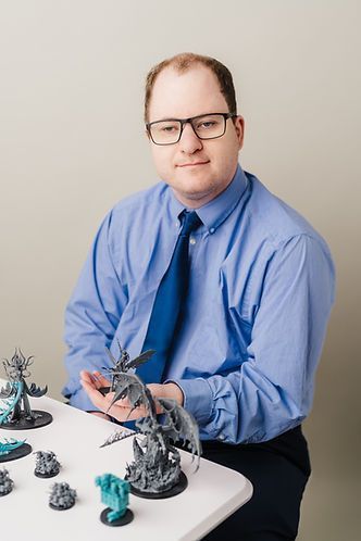 Man with glasses and blue shirt sits at a table with miniature fantasy figures. He is smiling and looking at the camera.