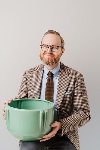 Man with glasses and a brown blazer holding a large, green bowl, smiling in front of a light gray background.