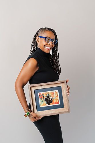 Smiling Black woman with locs and glasses holds a framed artwork. She wears a black outfit against a gray background.