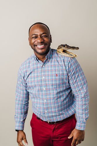Man in a blue plaid shirt and red pants smiles with a small alligator resting on his shoulder.