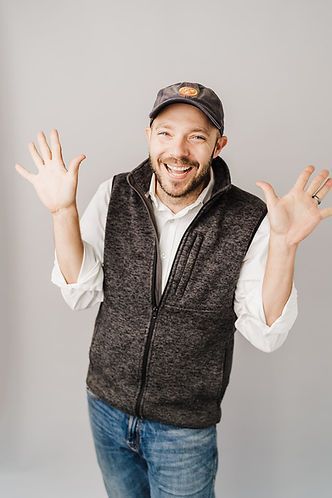 Man wearing a baseball cap, vest, and jeans, smiling with hands raised. Gray background.