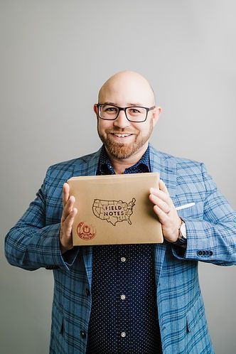 Man with glasses, in a blue blazer, holds a brown box with a US map, smiling against a grey background.