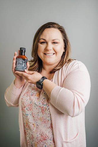Woman holding a bottle of sauce, smiling at the camera. She's wearing a floral shirt, pink sweater, and a watch. Gray background.