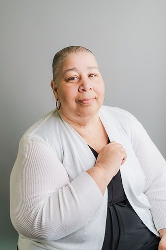An older Black woman in a white sweater and black top smiles while gesturing toward her chest. Gray background.