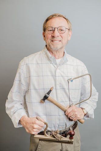 Man with glasses, holding a hammer, saw, and tools in a light gray studio background. He smiles at the camera.