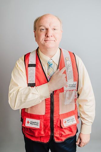 Man wearing a red safety vest with Red Cross logos, pointing to a badge, against a gray background.