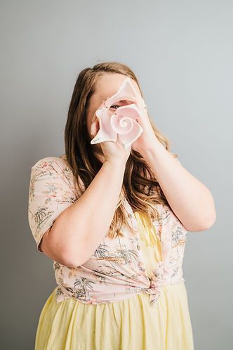 Woman holding a large pink conch shell in front of her face against a gray background. She wears a floral shirt and yellow dress.