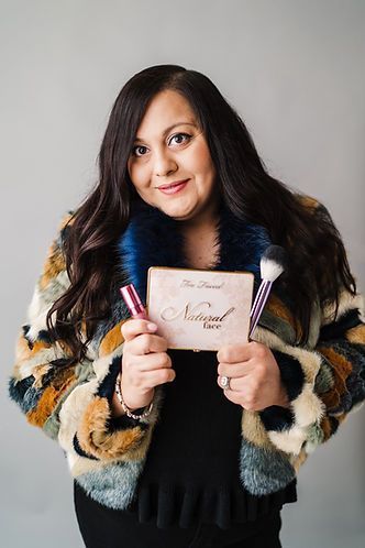 Woman holding Too Faced eyeshadow palette, makeup brush, and lipstick, wearing a patterned fur coat; smiling.
