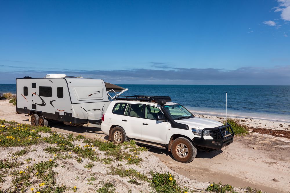 White SUV towing a gray caravan on a sandy beach next to the ocean under a blue sky. — The Caravan Clinic in Gold Coast, NSW