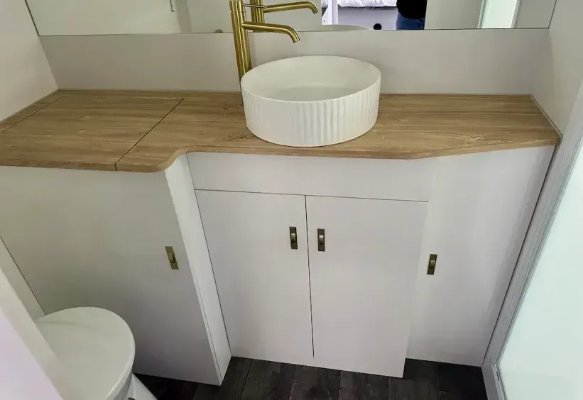Bathroom with a white vessel sink, gold faucet, and white cabinets with a wooden countertop. — The Caravan Clinic in Ballina, NSW