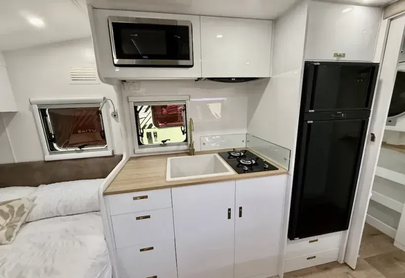 White caravan kitchen with microwave, fridge, sink, and stovetop; near a window and bed. — The Caravan Clinic in Tweed Heads, NSW