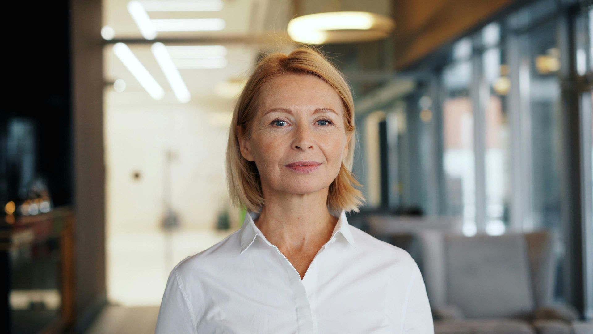 A woman in a white shirt is standing in a hallway and smiling at the camera.