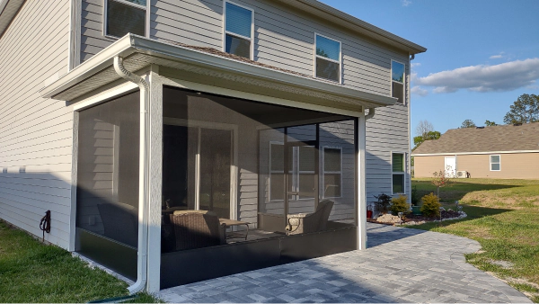 Screened-in porch with white framing attached to a two-story house with light gray siding. The porch has a gray paver patio.