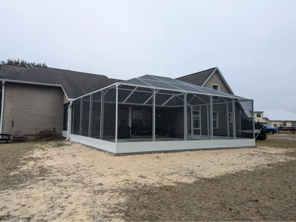 Screened-in patio attached to a house with a gray roof, beige siding, and a sandy yard.