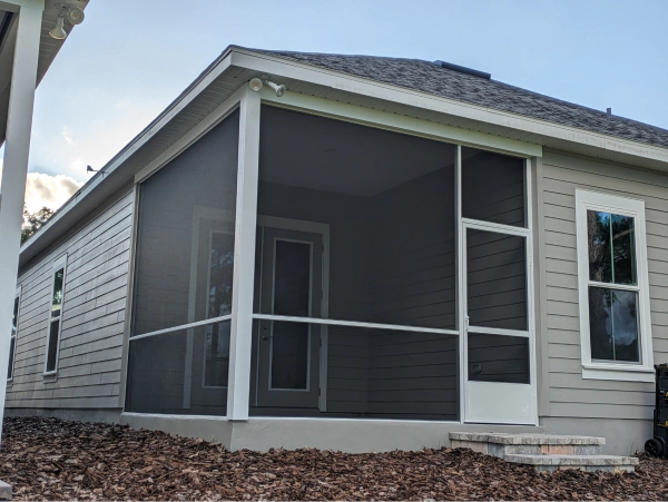 Screened-in porch with white trim, dark screen, and a door, attached to a light-colored house with a window.