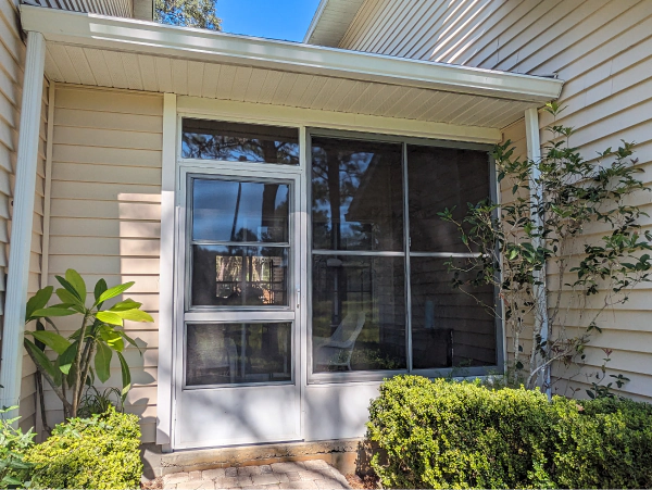 A white framed window with a dark screen, set in a light-colored siding wall, has plants in front of it.