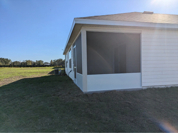 A white screened-in porch attached to a house on a sunny day, overlooking a grassy field.