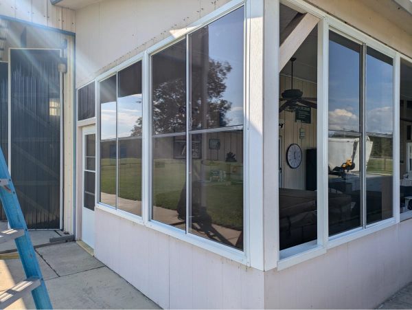 Sunroom with white frames and dark tinted windows. The room opens to a yard, with visible trees and sky. A ladder leans against the outside.