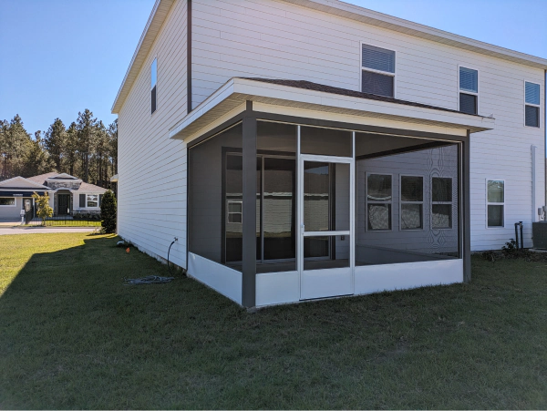 Screened-in porch attached to a two-story white house on a sunny day.  Gray frames enclose screens with windows. Green grass surrounds it.
