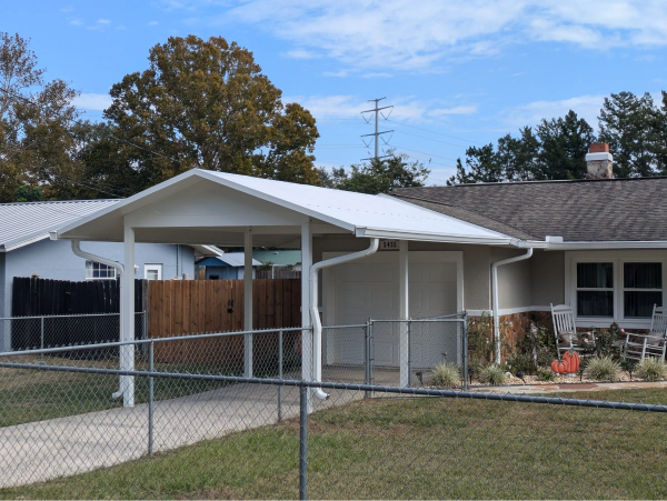 A white carport attached to a tan house. The carport has a white roof and supports, and a fence encloses the driveway.
