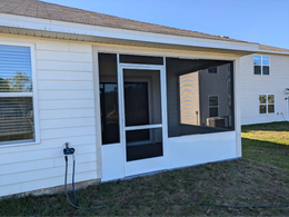 Screened-in porch attached to a white house, with a door and windows visible. The porch is white and has a dark screen.