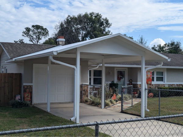 A white carport attached to a single-story house with a gray roof, driveway, and a chain-link fence.