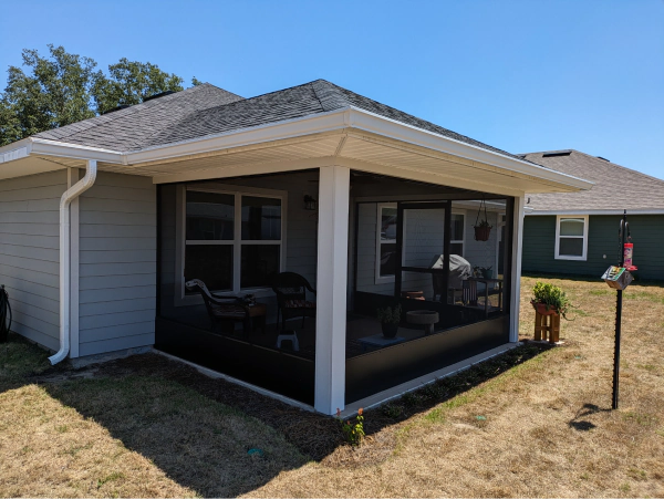 Screened-in patio attached to a house with gray siding, a dark roof, and a clear blue sky.