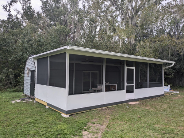 A white screened-in porch with dark screens and a gray roof, set in a grassy backyard.  A small shed is attached to the side.