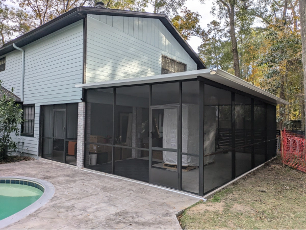 A screened-in porch attached to a light blue house, with a pool in the foreground and trees in the background.