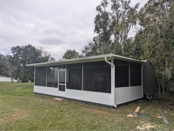 Screened-in white patio structure with dark screens and a white roof, situated on a grassy lawn. Trees are in the background.