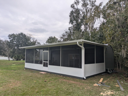 A white screened-in porch with a gray roof sits on a grassy yard, surrounded by trees under a cloudy sky.
