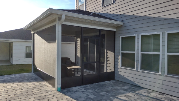 Screened-in patio attached to a house with grey siding. A brick patio floor and white trim surround the structure.