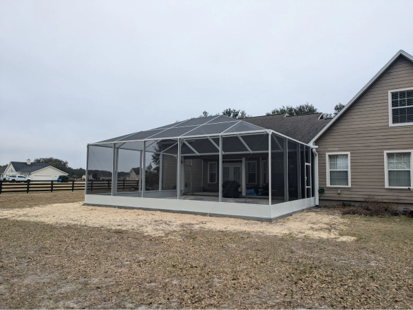 A screened-in patio attached to a beige house. The patio has a gray roof, white trim, and sits on a concrete base surrounded by gravel and grass.