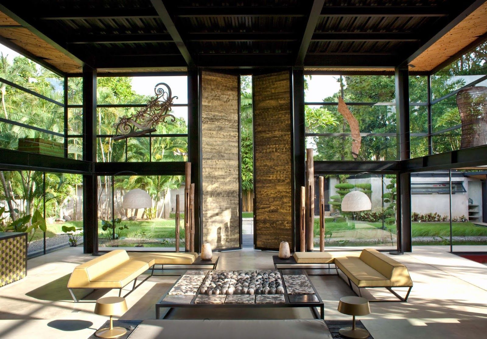 Salon ouvert avec baies vitrées, tons neutres et vue sur une végétation. Canapés, grand tapis et colonnes décoratives.