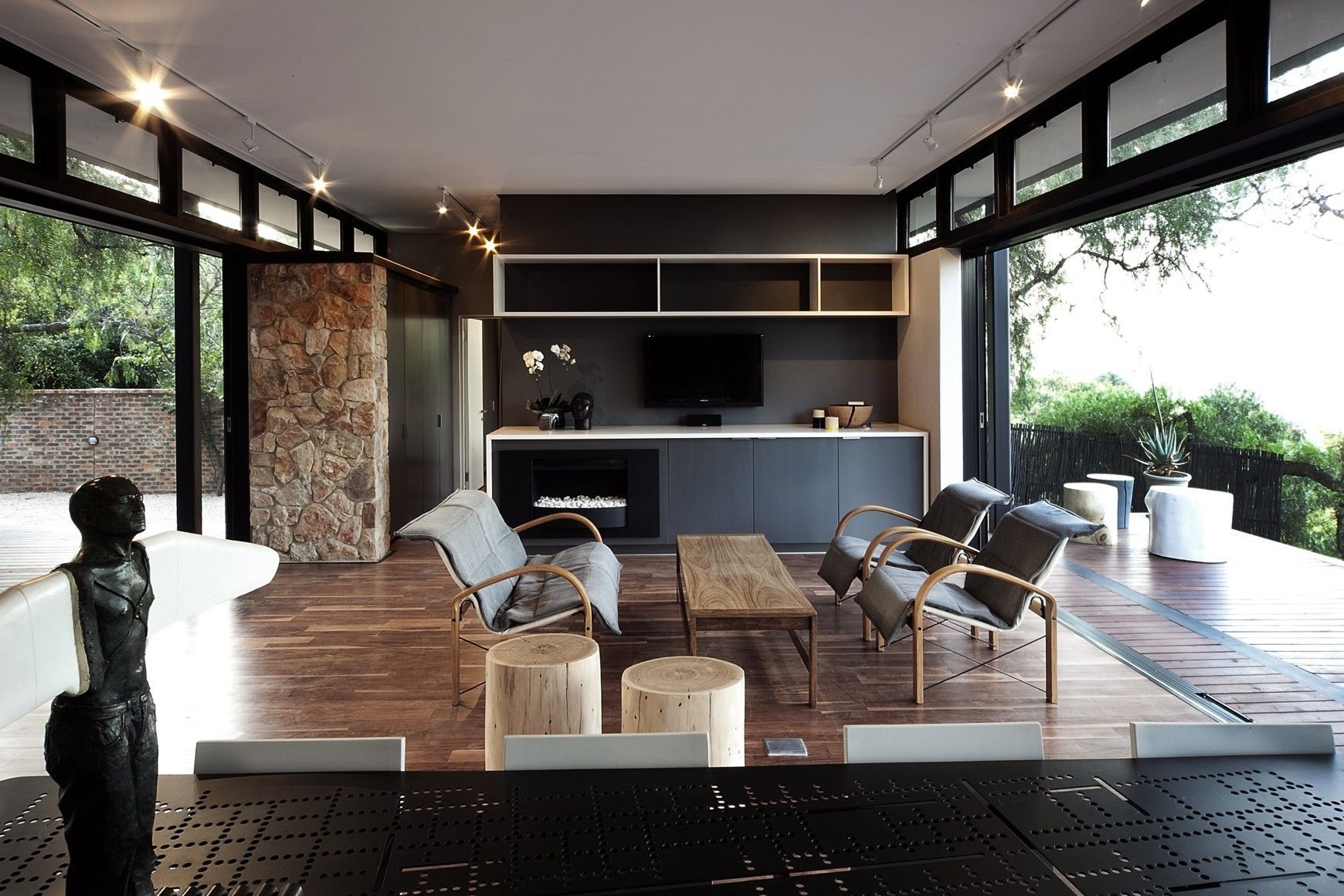 Salon moderne et spacieux avec murs en verre ouverts, armoires gris foncé, parquet et sièges donnant sur la verdure.