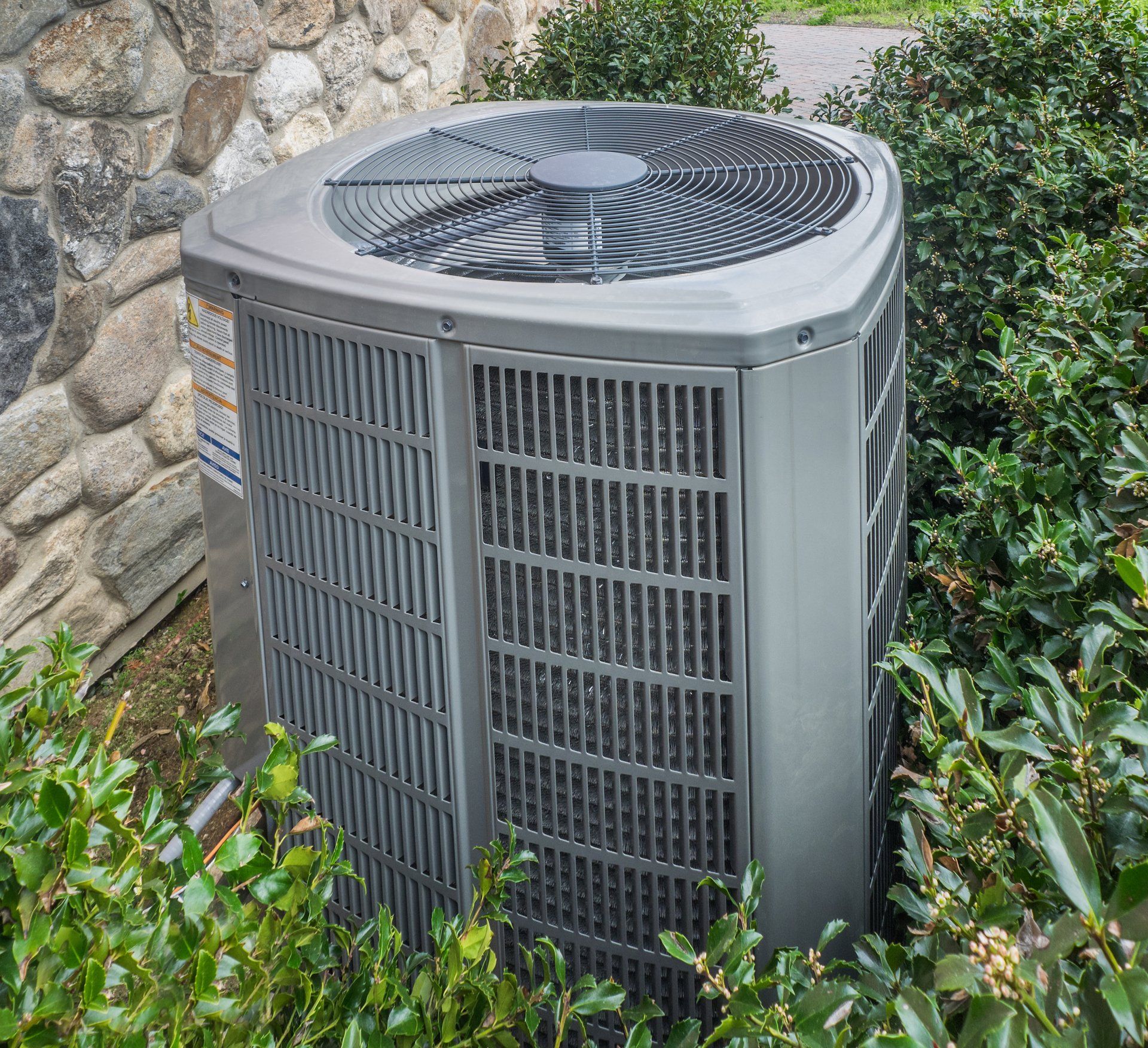 A large air conditioner is sitting in a bush next to a stone wall.