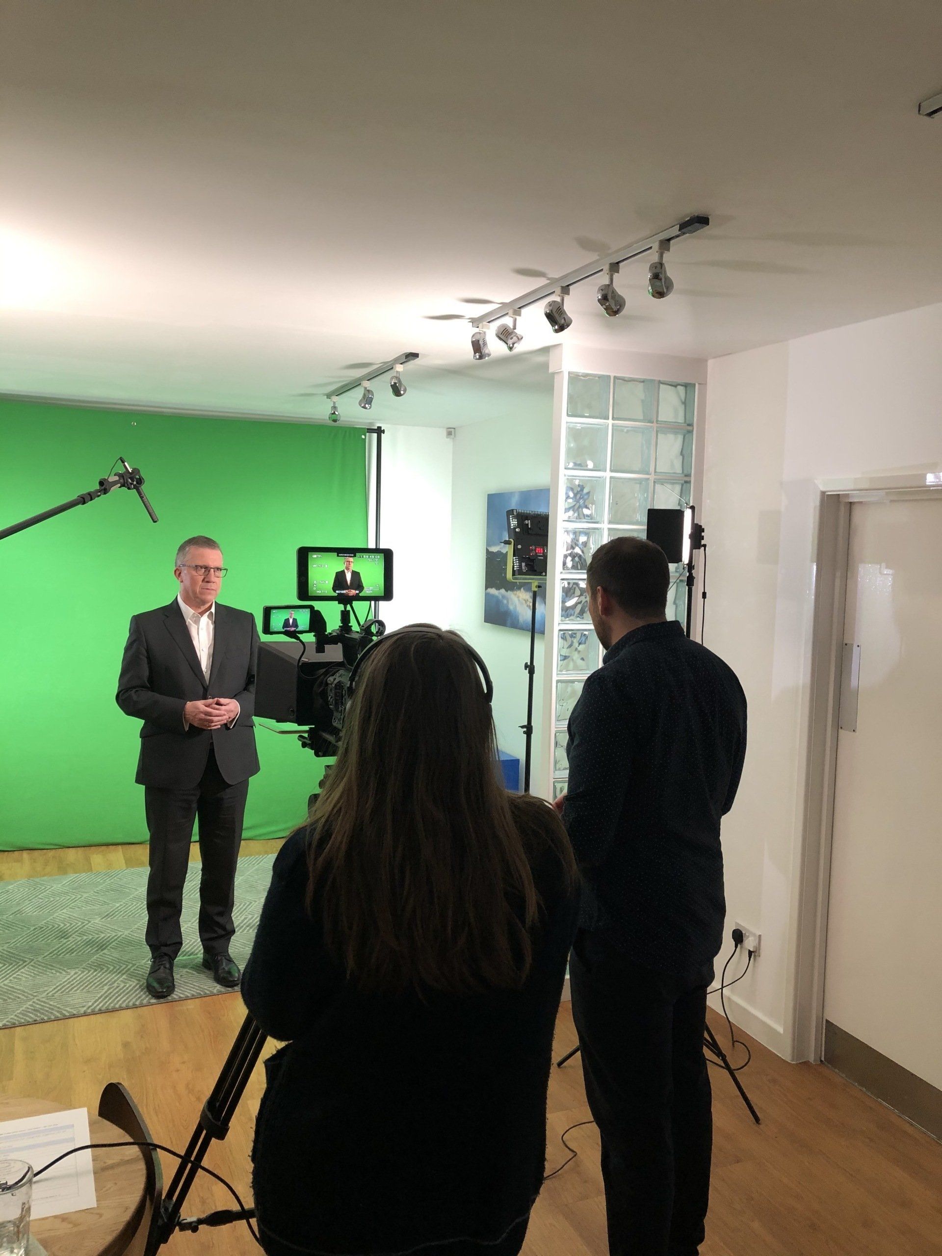 A man in a suit is standing in front of a green screen in a studio.