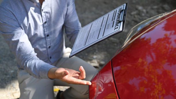 A man is kneeling down next to a red car holding a clipboard.