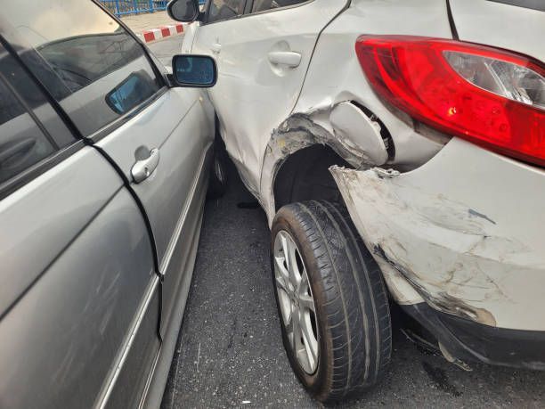 A white car with a damaged fender is parked next to a silver car.