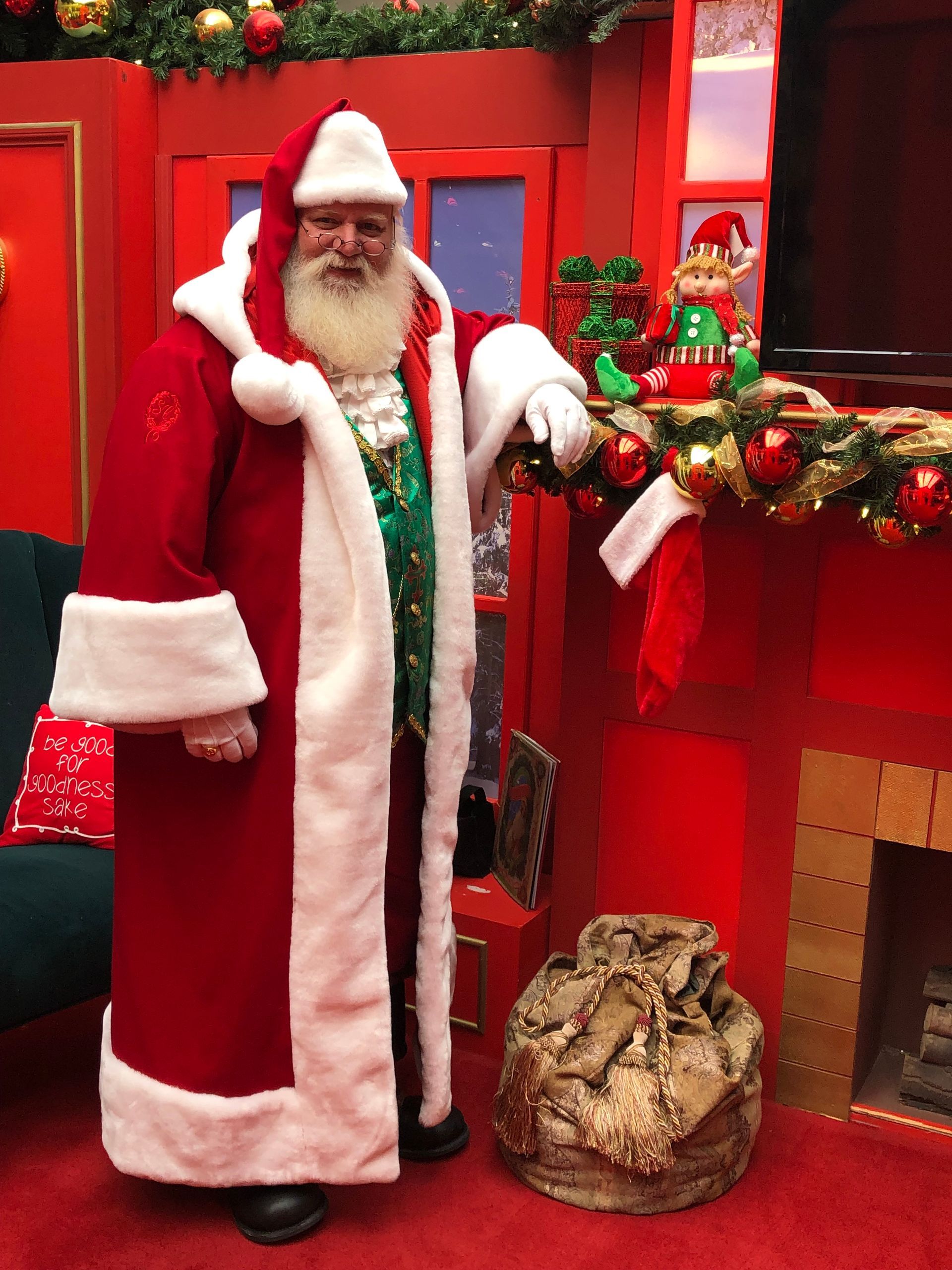 Santa Claus in a red suit stands by a fireplace with a sack of gifts.
